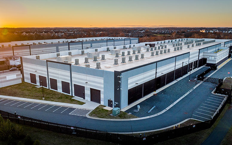 Aerial view of a large data center at sunset. The building is surrounded by a paved parking lot. The sun sets casting warm colors in the sky.