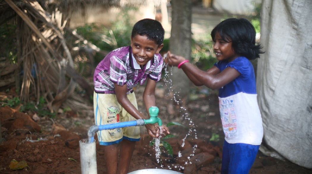 Two children wash their hands and play with water from a ground faucet. 