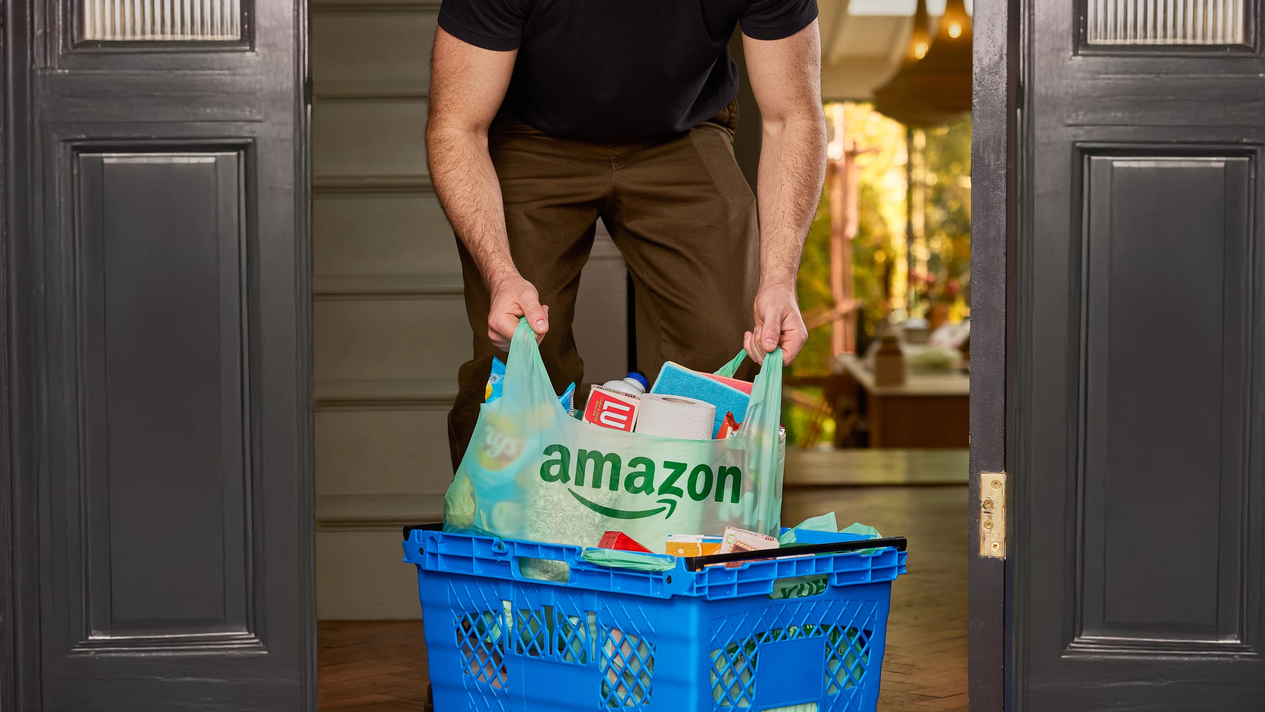 A person lifts groceries out of a blue crate in a green bio bag with the Amazon logo.