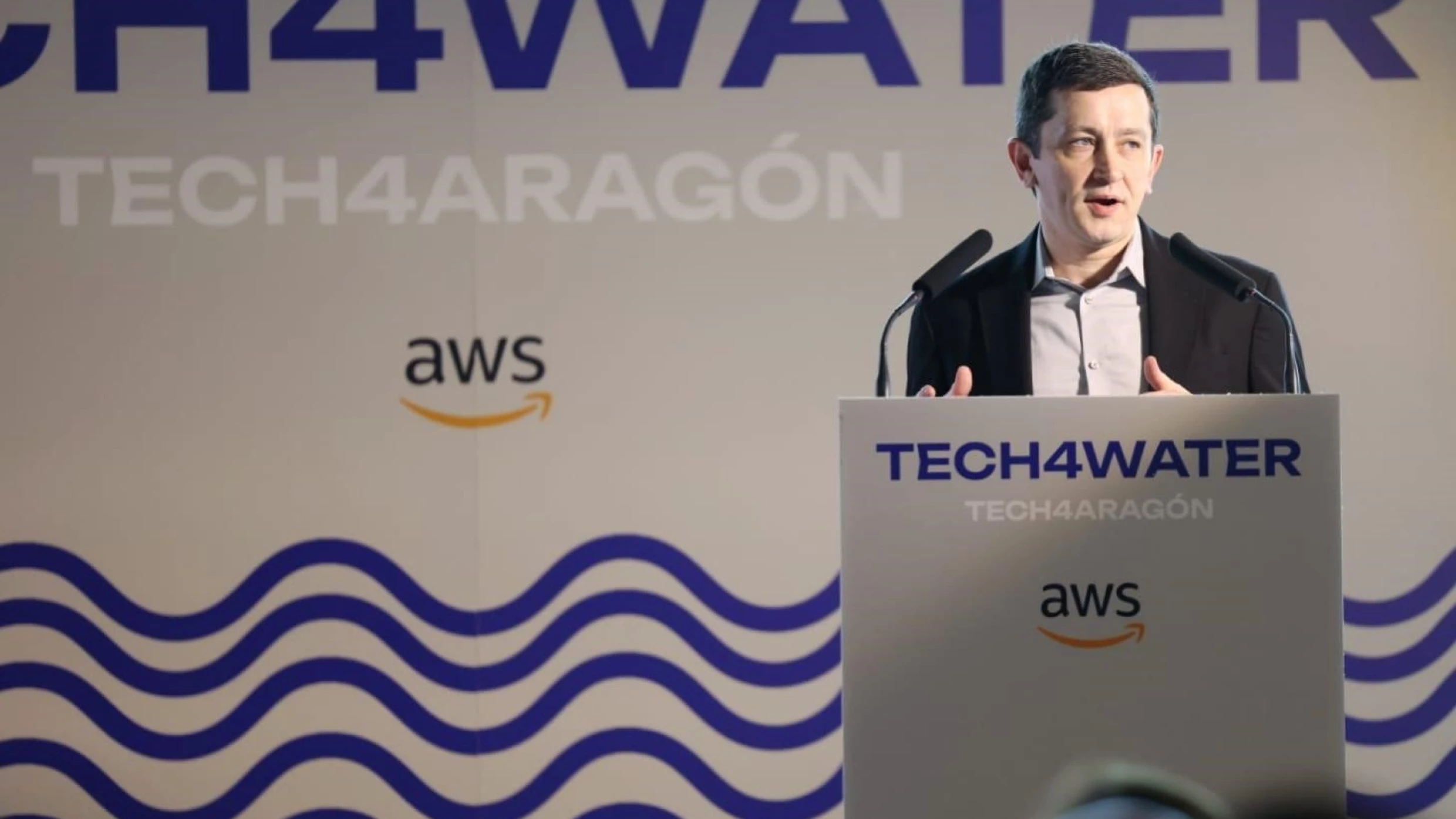 A person stands at a speaker podium in front of a wall that says "Tech4Aragon" with an AWS logo. 