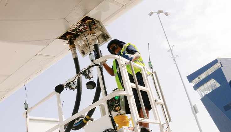 A person wearing safety gear is shown operating machinery that provides fuel to a large aircraft beneath text about "First mile" transportation. 