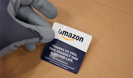 A gloved hand is shown placing a sticker with the Amazon logo on a cardboard box. 