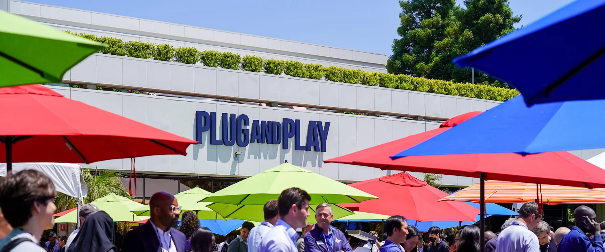 A group of people outside under umbrellas by a building that says "Plug and Play."