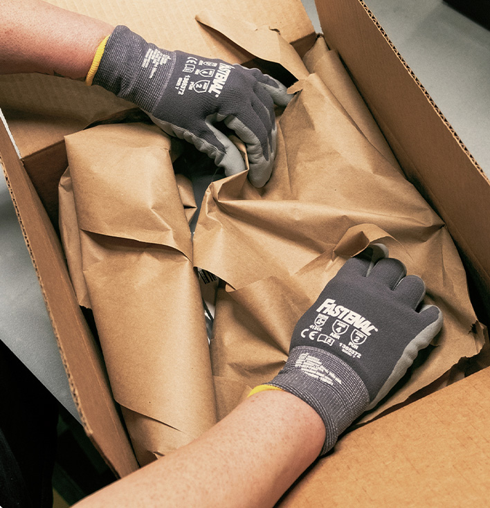 A person wearing gloves unpacks a box filled with brown paper. 
