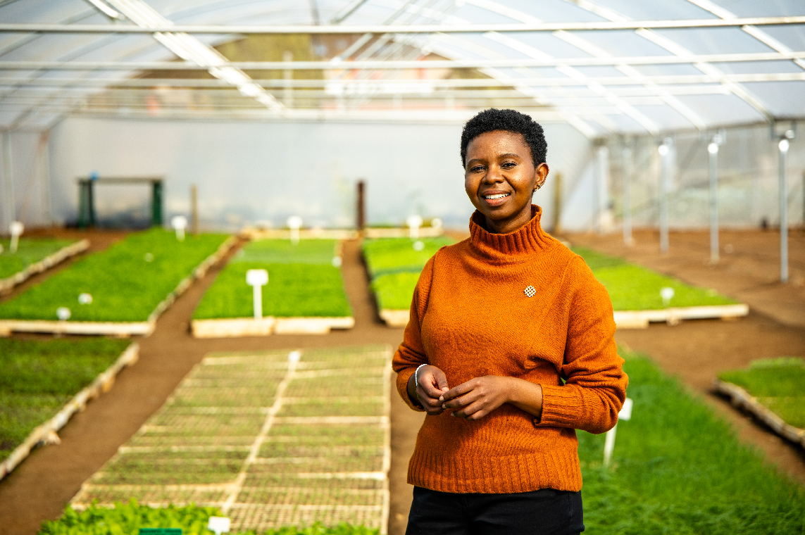 A person stands smiling in front of plants in a greenhouse. 