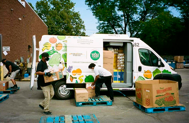 Two people unload boxes of supplies from a Whole Foods Market branded van.