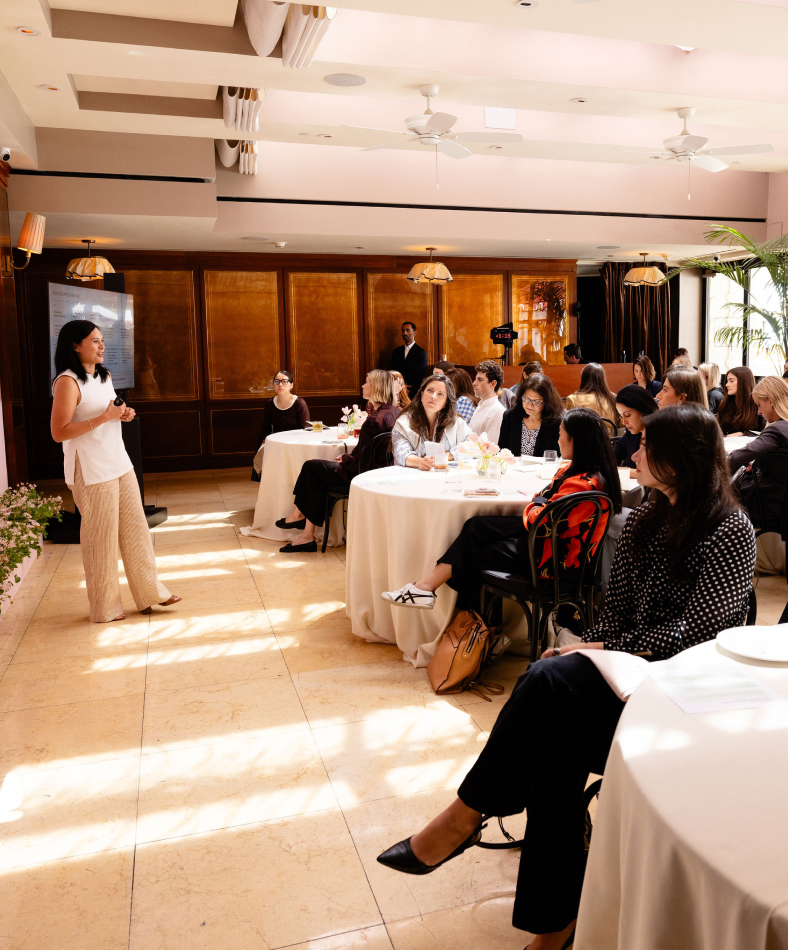 A woman gives a presentation to a seated audience at a daytime networking event.