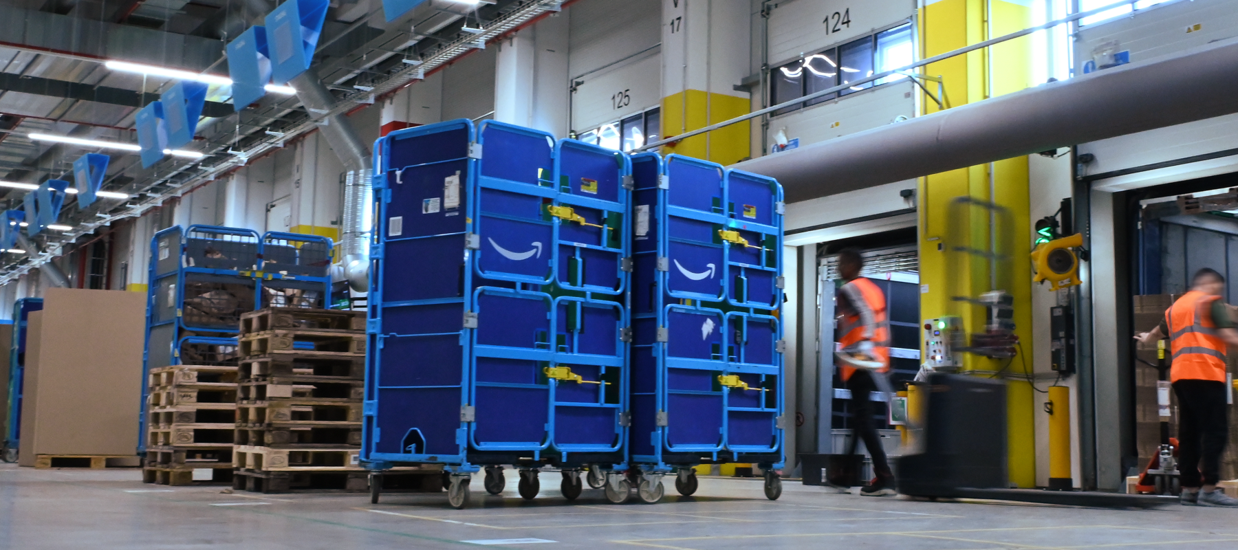 Two blue carts are shown in a warehouse near a loading dock, with two people in orange vests working nearby. 
