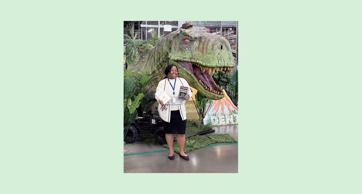 A person stands and smiles in front of a dinosaur sculpture. 