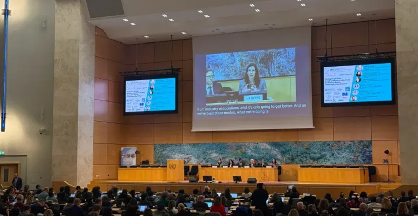 A person shown on a screen speaking from a podium to a crowd of people. 