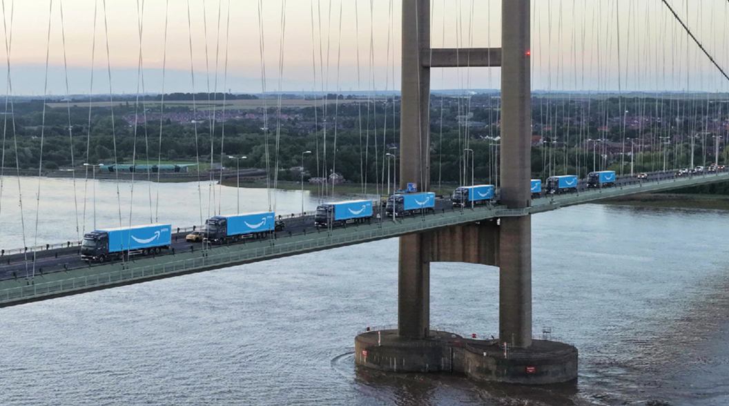 A line of long-haul delivery vehicles drives across a bridge over water. 