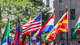 Country flags stand in a row with tall buildings in the background. 