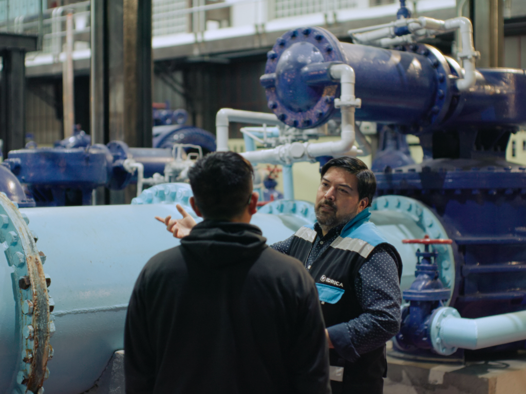 Two people talk while standing in front of equipment holding water. 