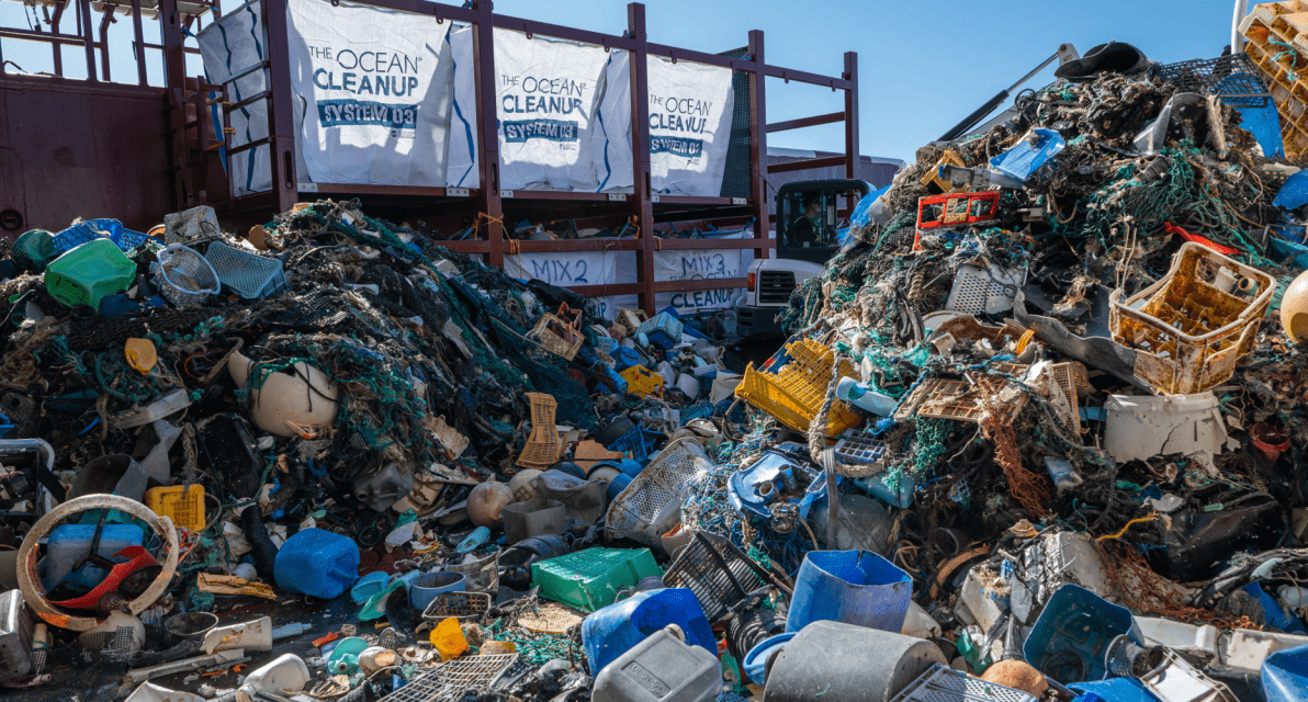 Piles of trash gather in front of bags labeled with The Ocean Cleanup logo. 