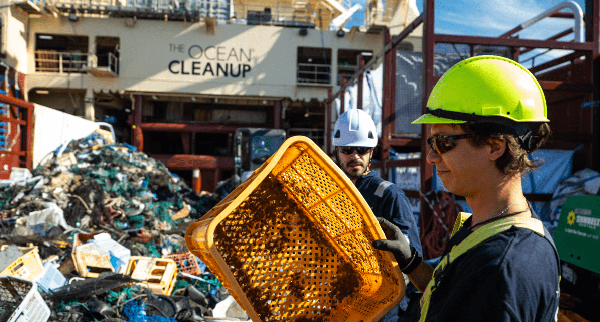 A person wearing a hard hat holds up an orange bin used to sort trash. 