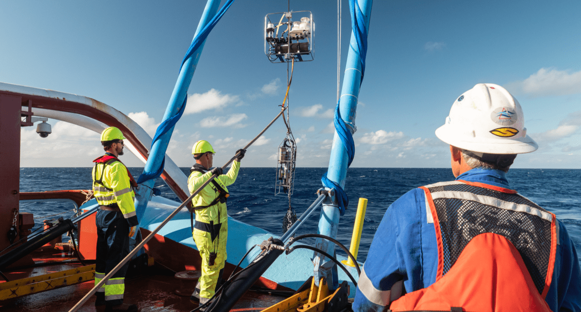 Three people wearing safety gear guide a piece of machinery into the ocean from a boat. 