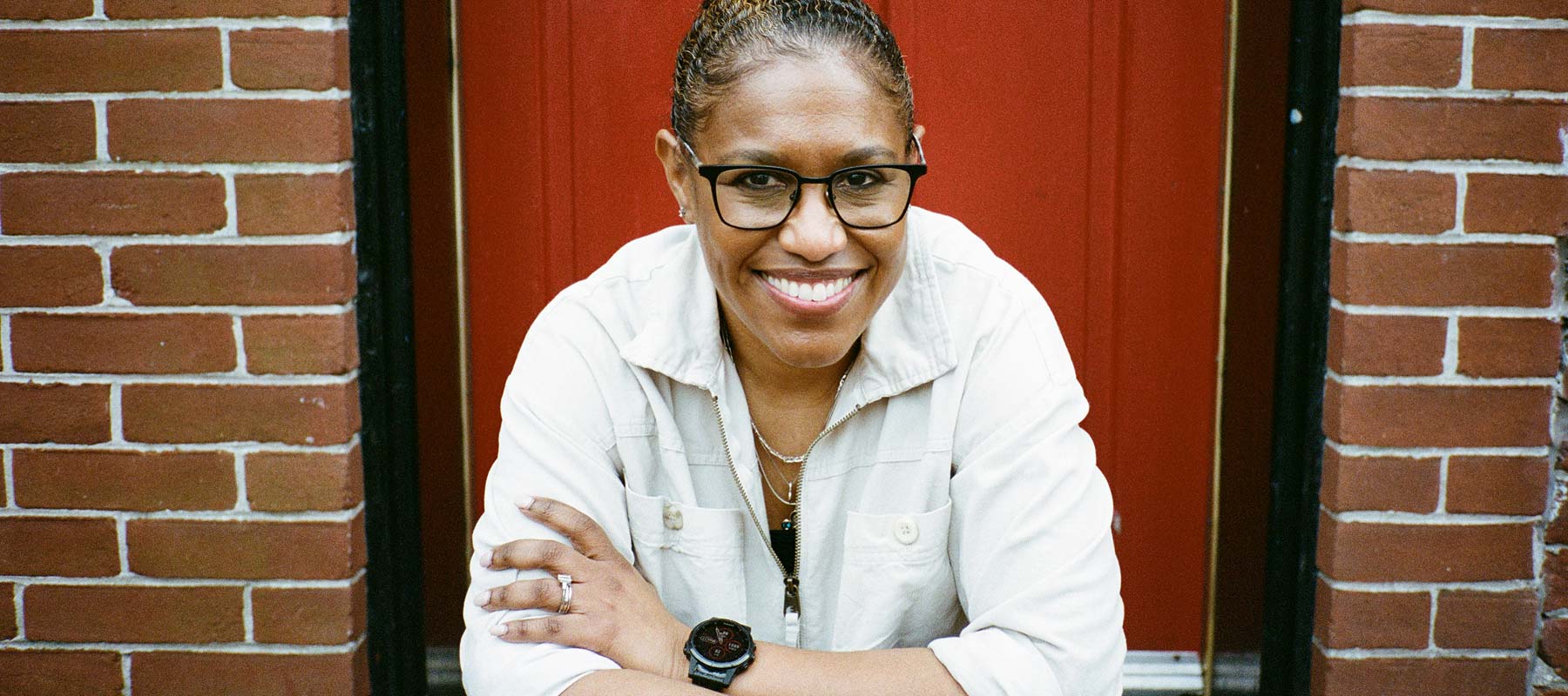 A person sits smiling in front of a brick building with a red door. 