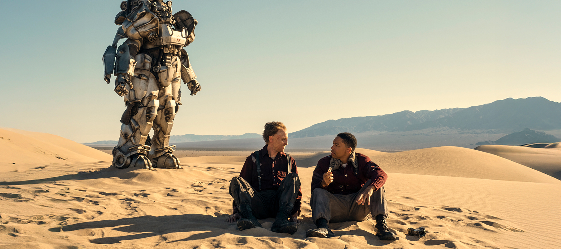 Two people sit in the sand with a robot in the background.