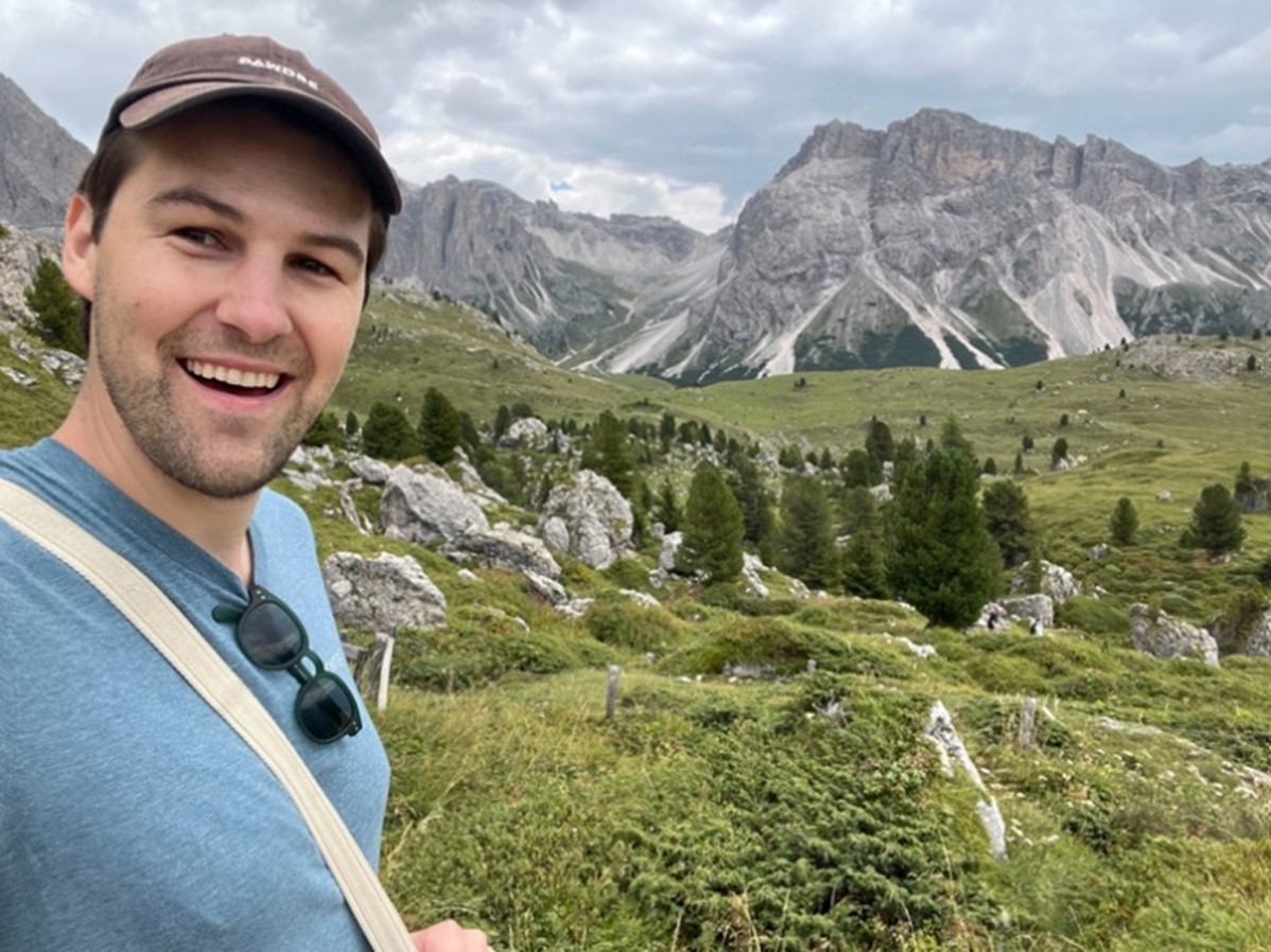 A person stands smiling in front of a mountain landscape. 
