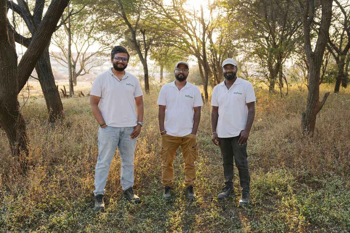 Three people stand smiling for a photo in a field surrounded by trees. 