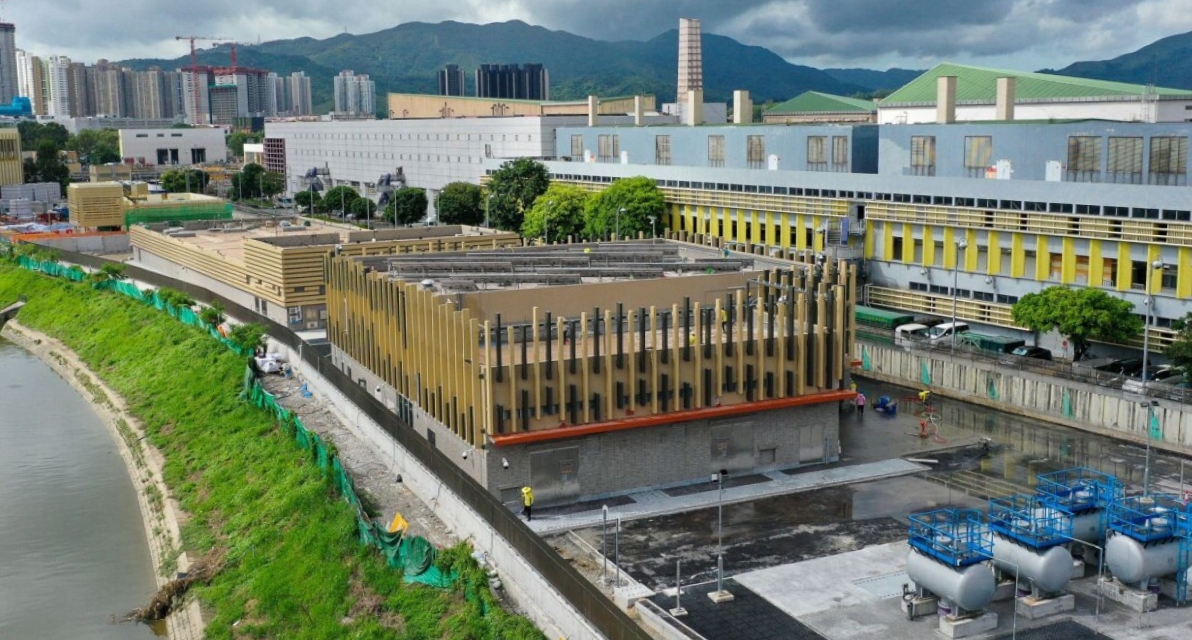 Buildings sit along a bank of water with lush mountains in the background. 